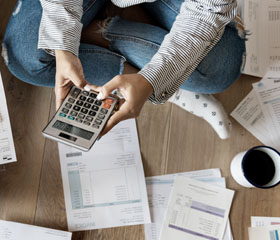 Una persona sentada en el suelo rodeada de facturas, revisando y sumando cuentas